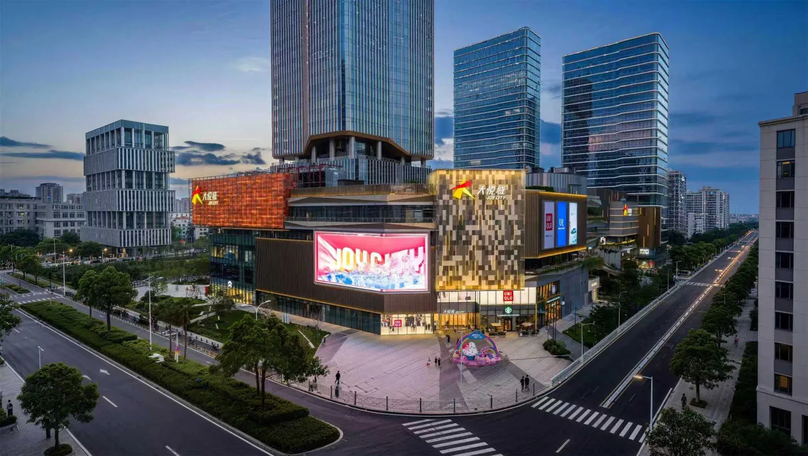 Sanya Joy City's lit facades and big screen at dusk, with office towers behind