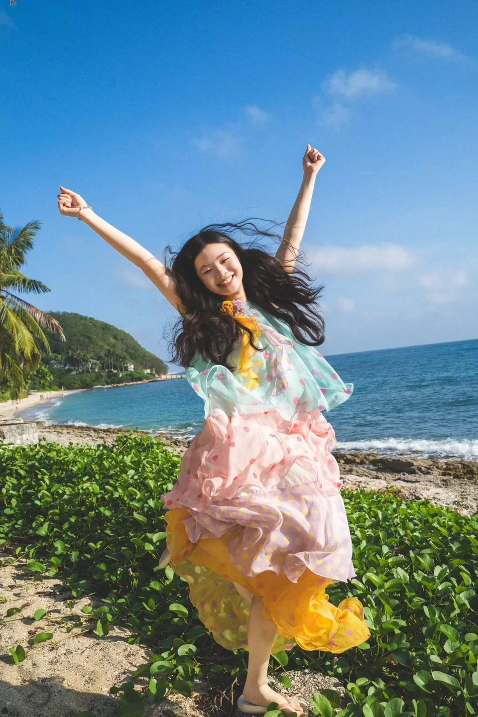 A woman in a colourful flowing dress beside a palm-lined coastline and blue sea