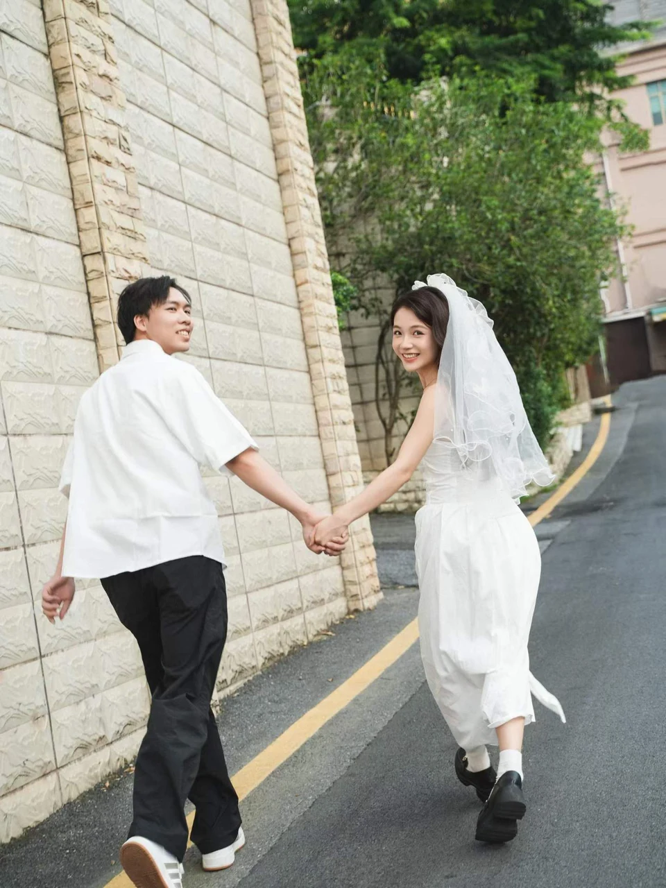 A couple in white holding hands on a street beside a stone wall and greenery