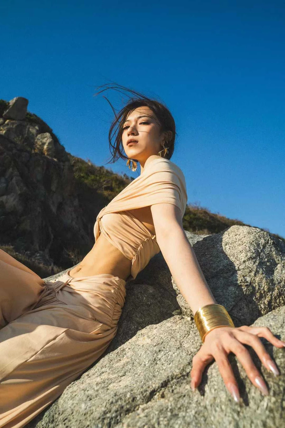 A woman in a beige outfit reclining on coastal rocks beneath a deep blue sky