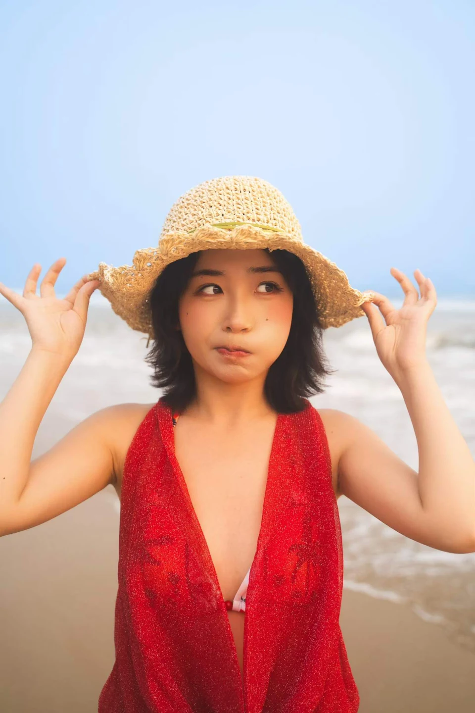 A woman in a red top holding the brim of a straw hat with the beach behind her
